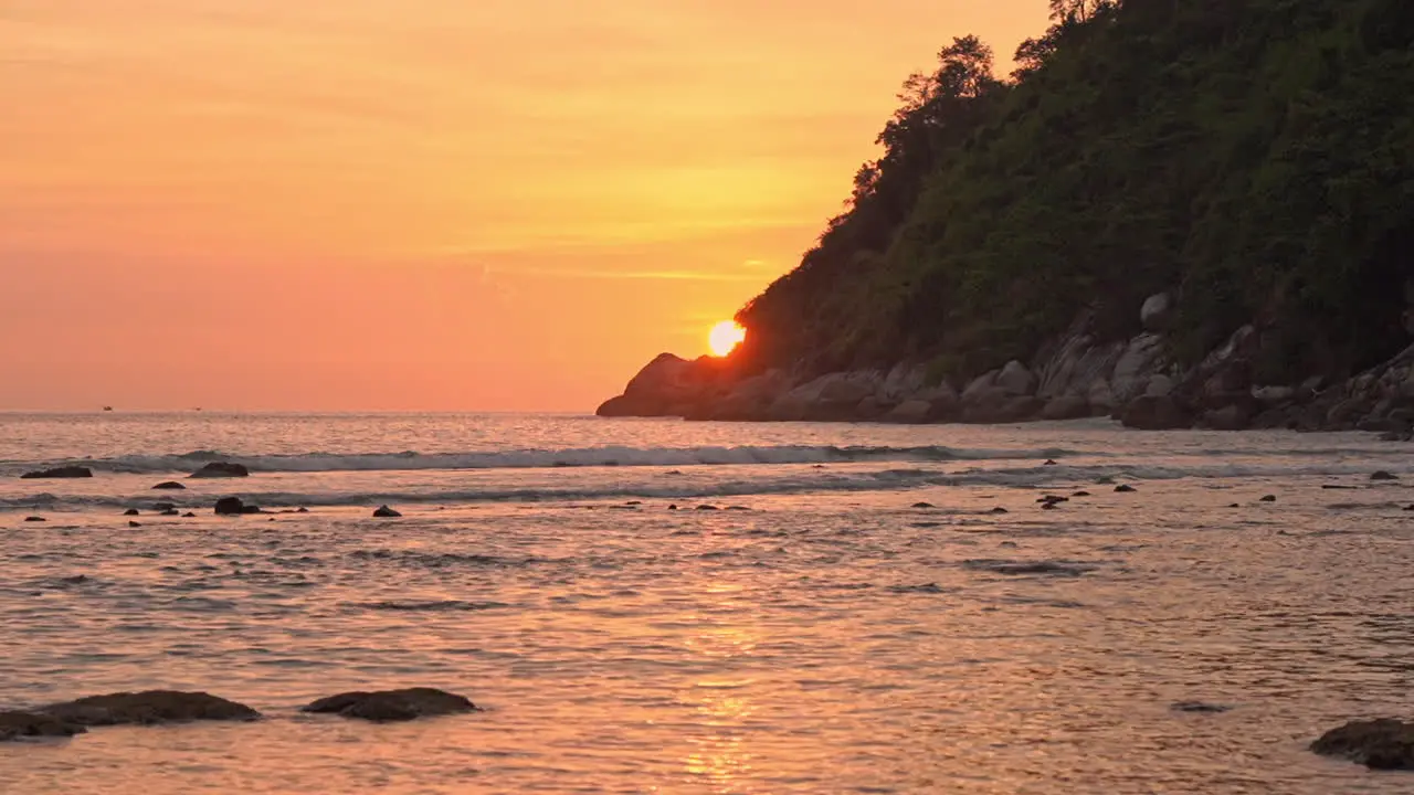 A dramatic orange sun dips behind a mountain along the coastline