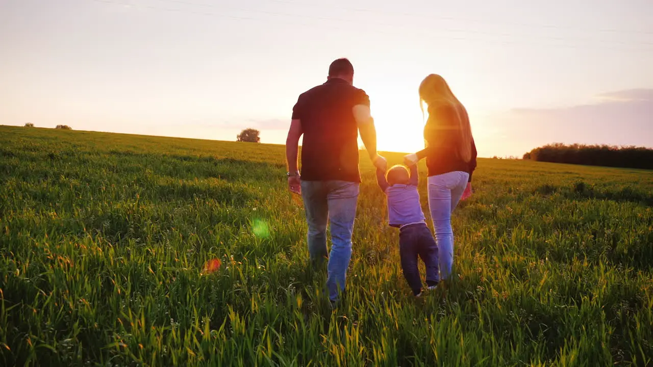 Happy Young Parents Are Walking Along The Field The Father Is Carrying The Baby Son In His Arms