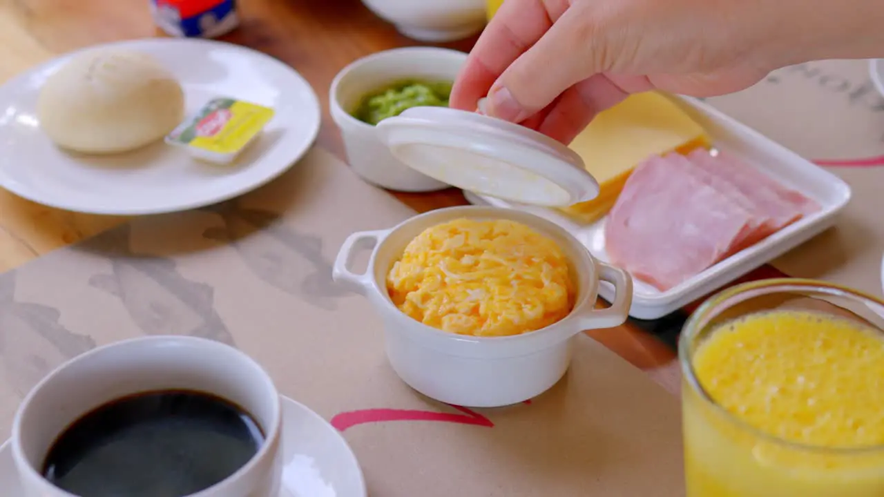 woman lifting the lid of a pot containing hot scrambled eggs next to a cup of coffee for breakfast