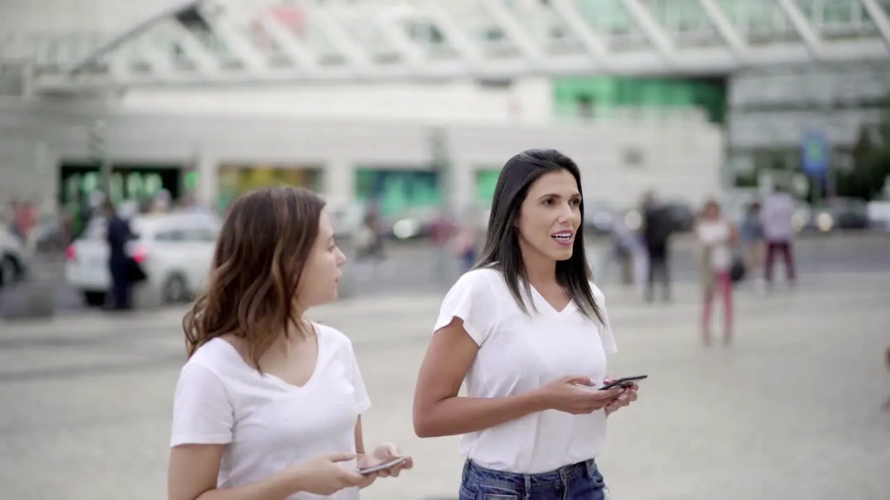 Two women walking on street with phones and talking