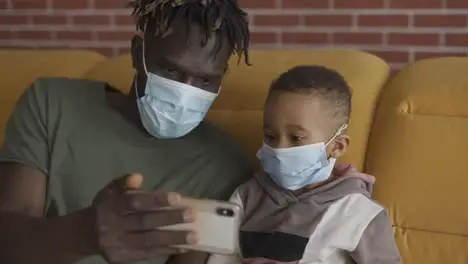 Father and son are sitting on a sofa in medical face masks watching video on smartphone