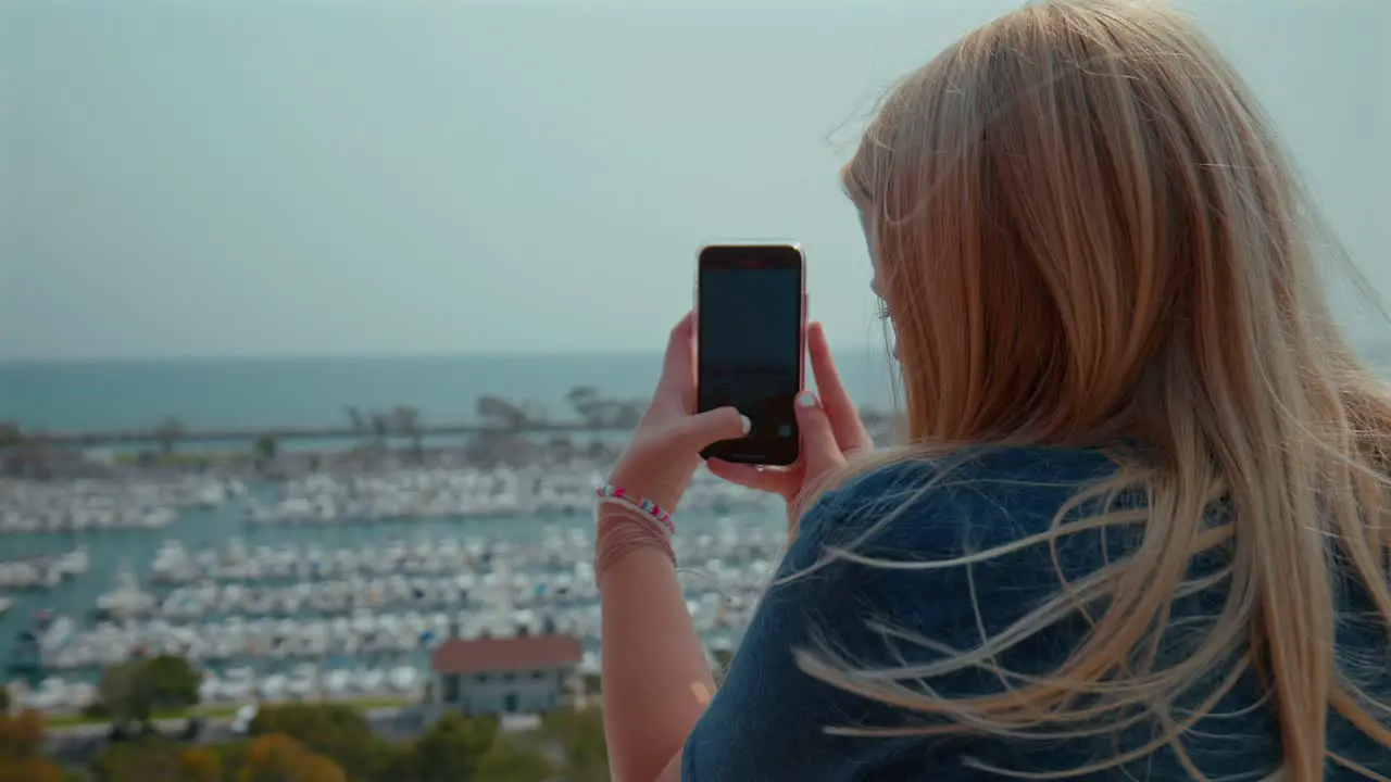 Blonde teenage caucasian girl taking pictures with her cell phone of a harbor in California