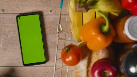 Overhead Studio Shot Of Person Using Green Screen Mobile Phone Next To Basic Food Items In Supermarket Wire Shopping Basket 2