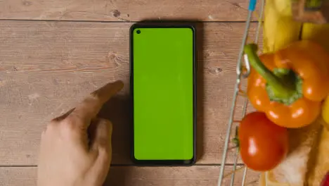 Overhead Studio Shot Of Person Using Green Screen Mobile Phone Next To Basic Food Items In Supermarket Wire Shopping Basket 1