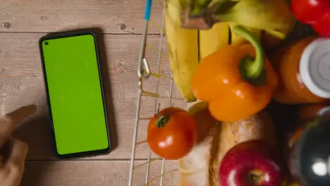 Overhead Studio Shot Of Person Using Green Screen Mobile Phone Next To Basic Food Items In Supermarket Wire Shopping Basket 3