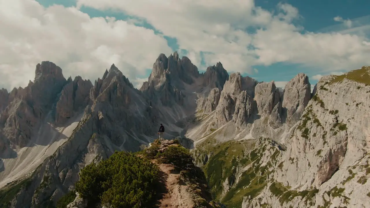 Parralax drone shot with lone tourist watching panoramic view with tall steep rocky mountains partly clouded sky in the background hiking in the Alps majestic landscape cinematic color grade