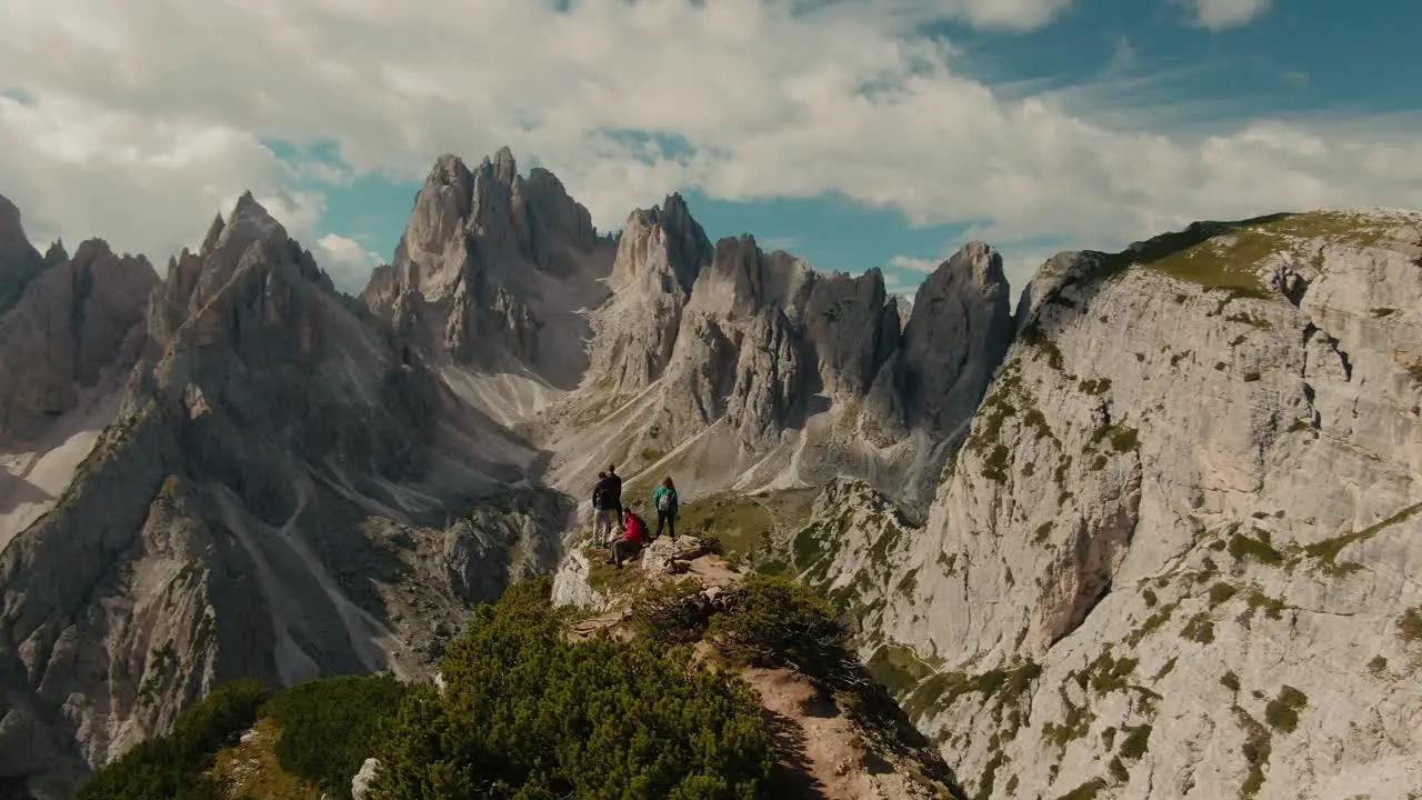 Parralax drone shot with tourists watching panoramic view with tall steep rocky mountains partly clouded sky in the background hiking in the Alps majestic landscape cinematic color grade