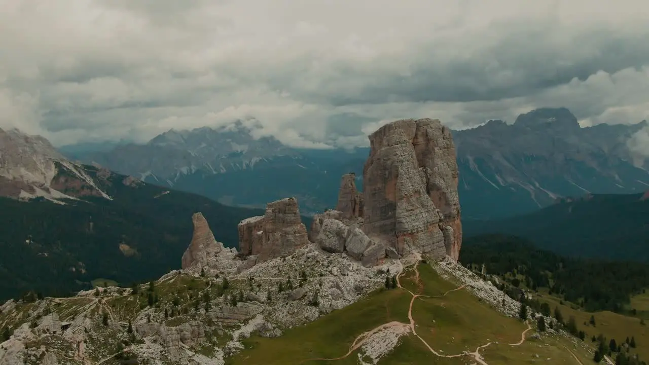 Aerial view aproaching massive rock formations with distant tall mountains in the background green forest and fields at the bottom cloudy day cinematic color grade