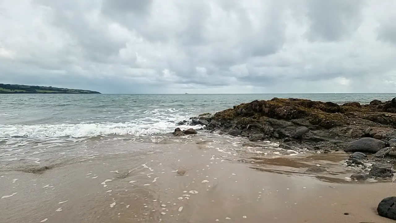 slow motion calming ocean waves washing across Anglesey island splashing over rocks on golden sandy beach overcast morning