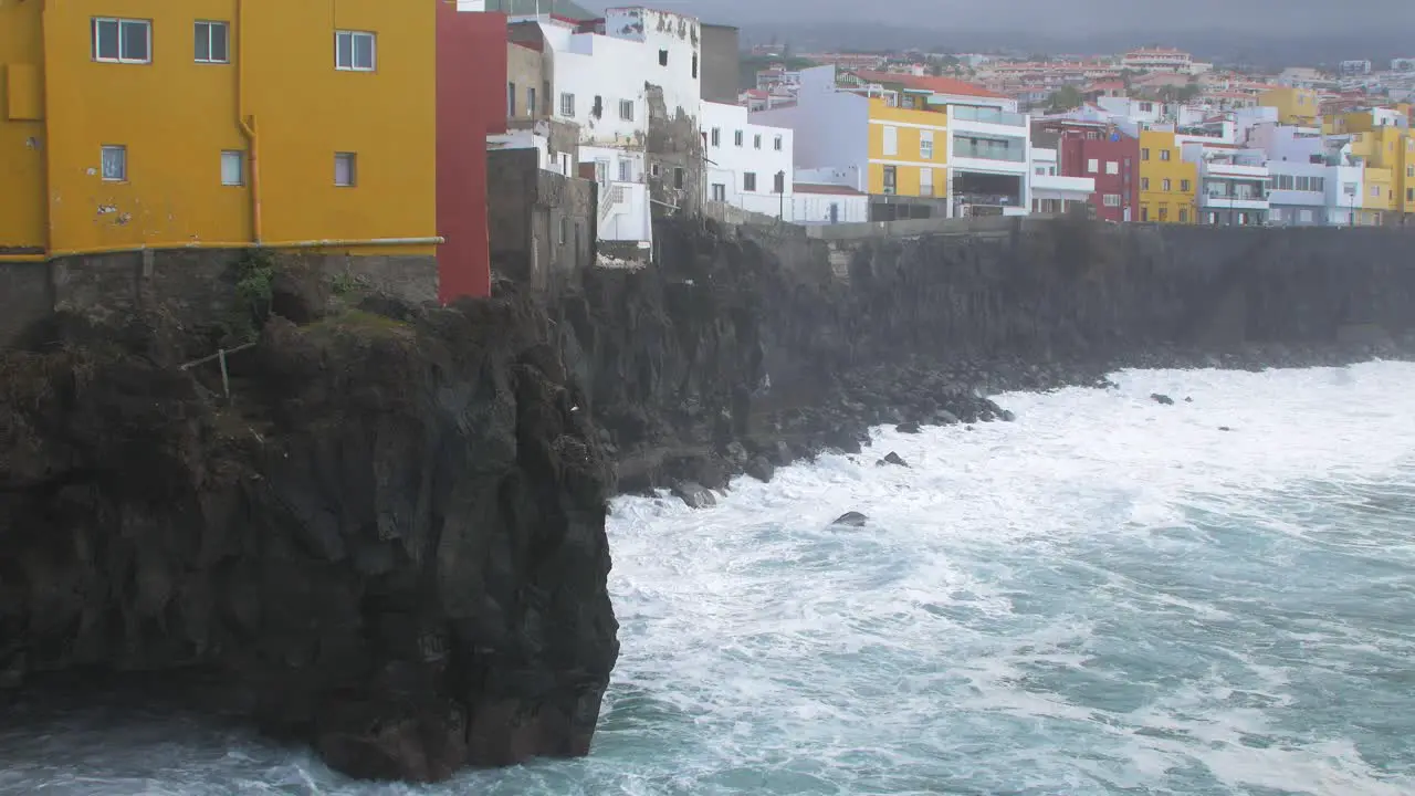 Big waves of the Atlantic Ocean breaks on a rocky coast on a sunny day during a storm in Puerto de la Cruz in the Canaries  distant colorful houses medium wide shot