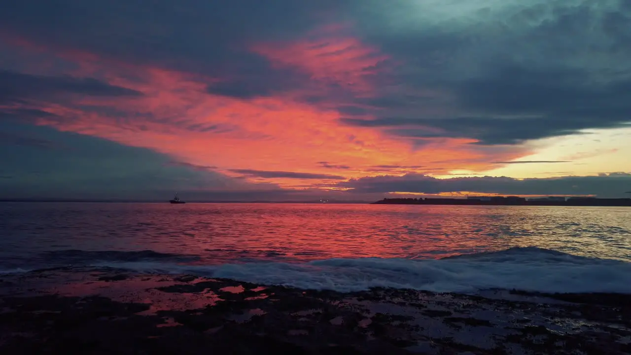 Burning red clouds during sunset over ocean water with wave breaking on shore