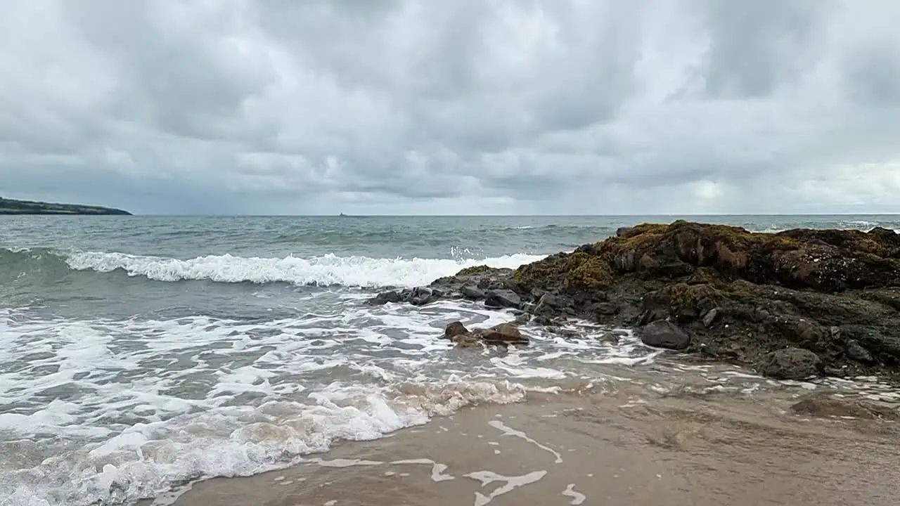 Slow motion ocean waves washing across Anglesey island splashing over rocks on golden sandy beach overcast morning