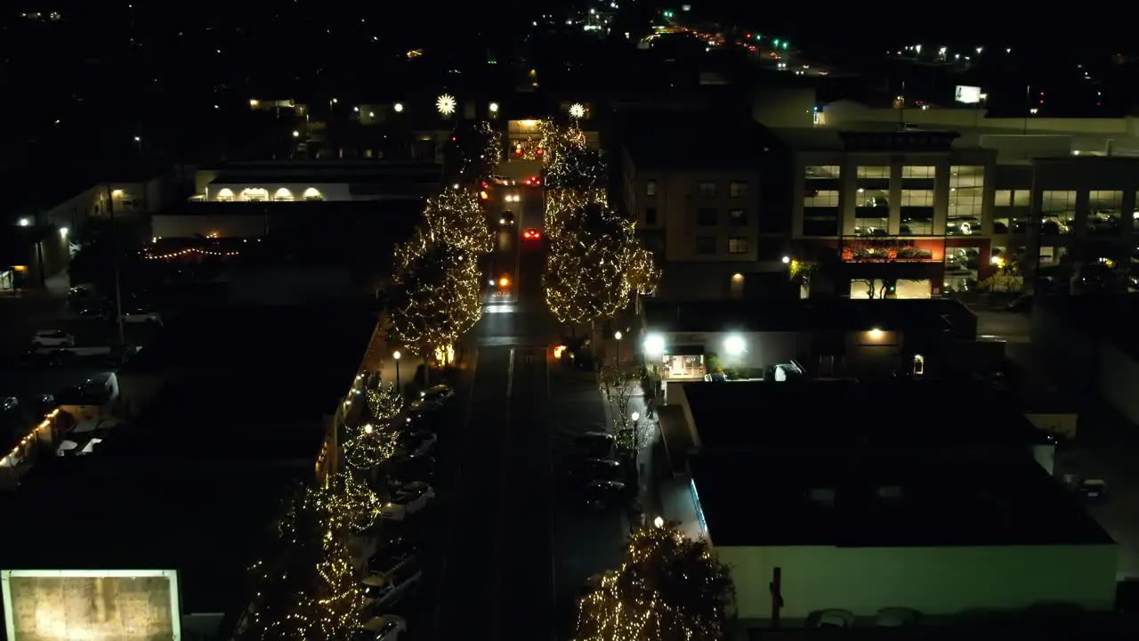 Small town during Holiday season of Christmas times street trees lit up with lights as cars drive down street at night aerial top down view