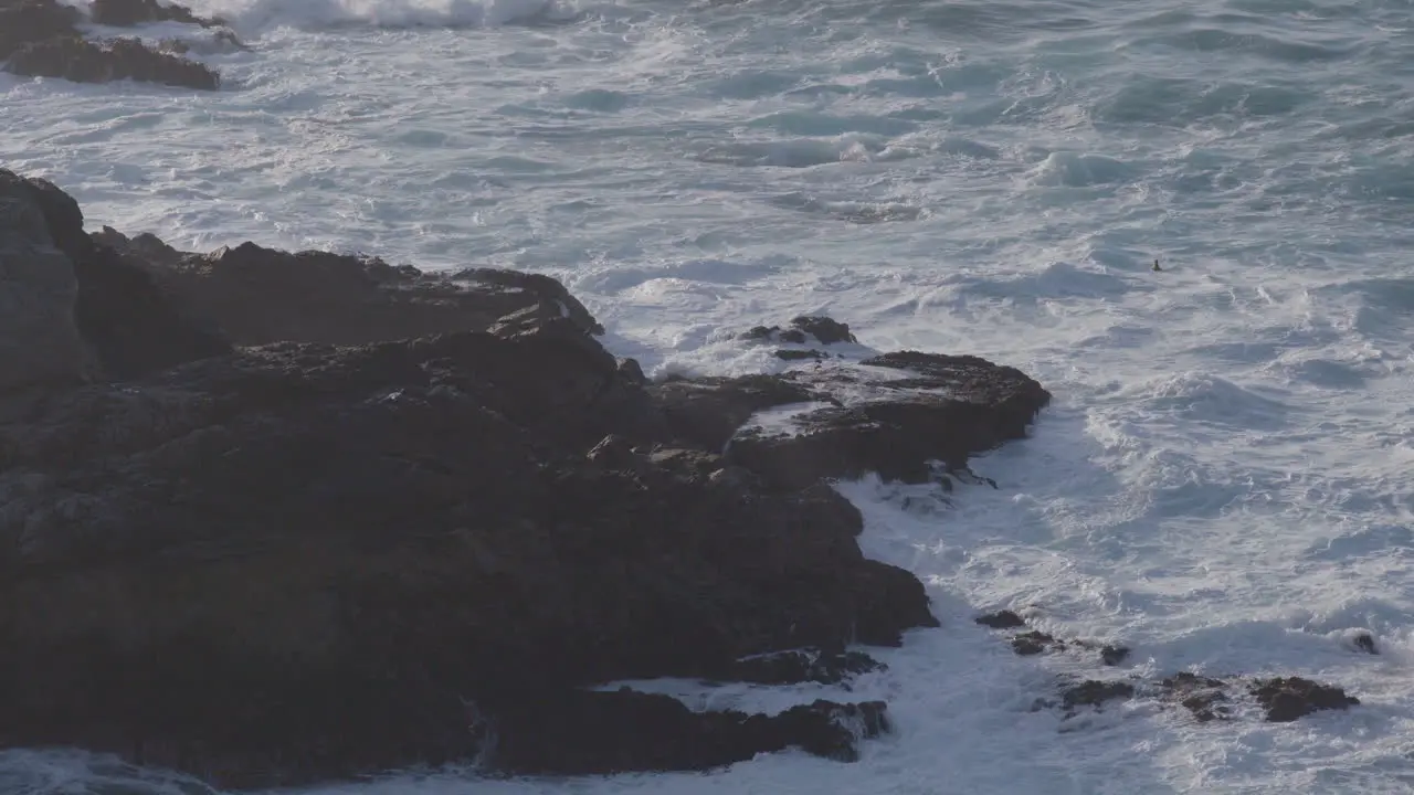 Stationary shot from above of waves crashing against giant boulders located in Big Sur California