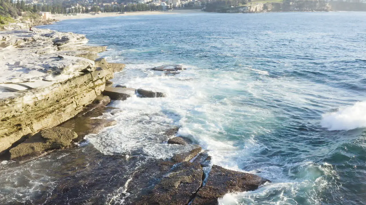 While fringed waves crashing onto ocean rocks POV drone moving slowly away from cliff face Coogee Beach Sydney Australia