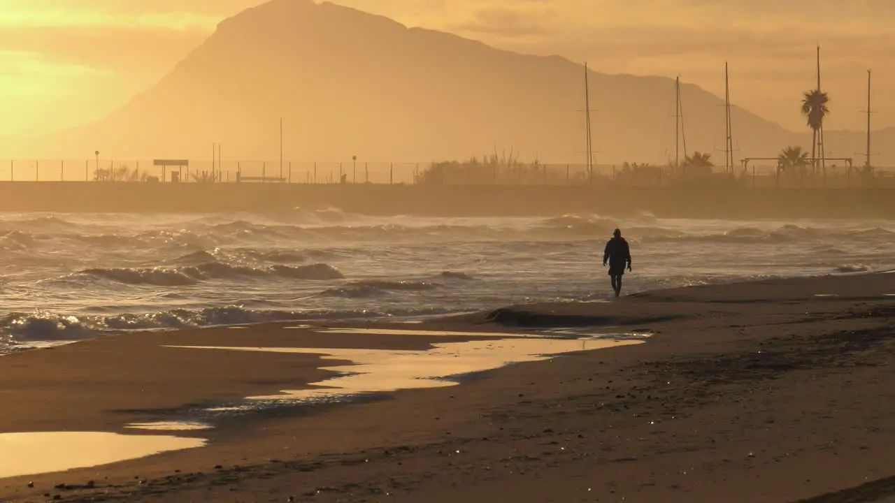 Sandy beach at sunrise with old man walking along shore mountain in background wide shot