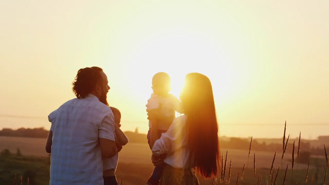 Young Man And Woman Playing With Young Children Tosses Them In The Air Against The Sunset Background