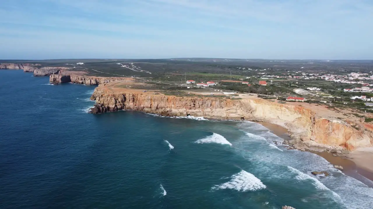 droneshot of the beautiful tonel beach with big waves rolling in the bay and sagres on the hills southern portugal with sunny weather