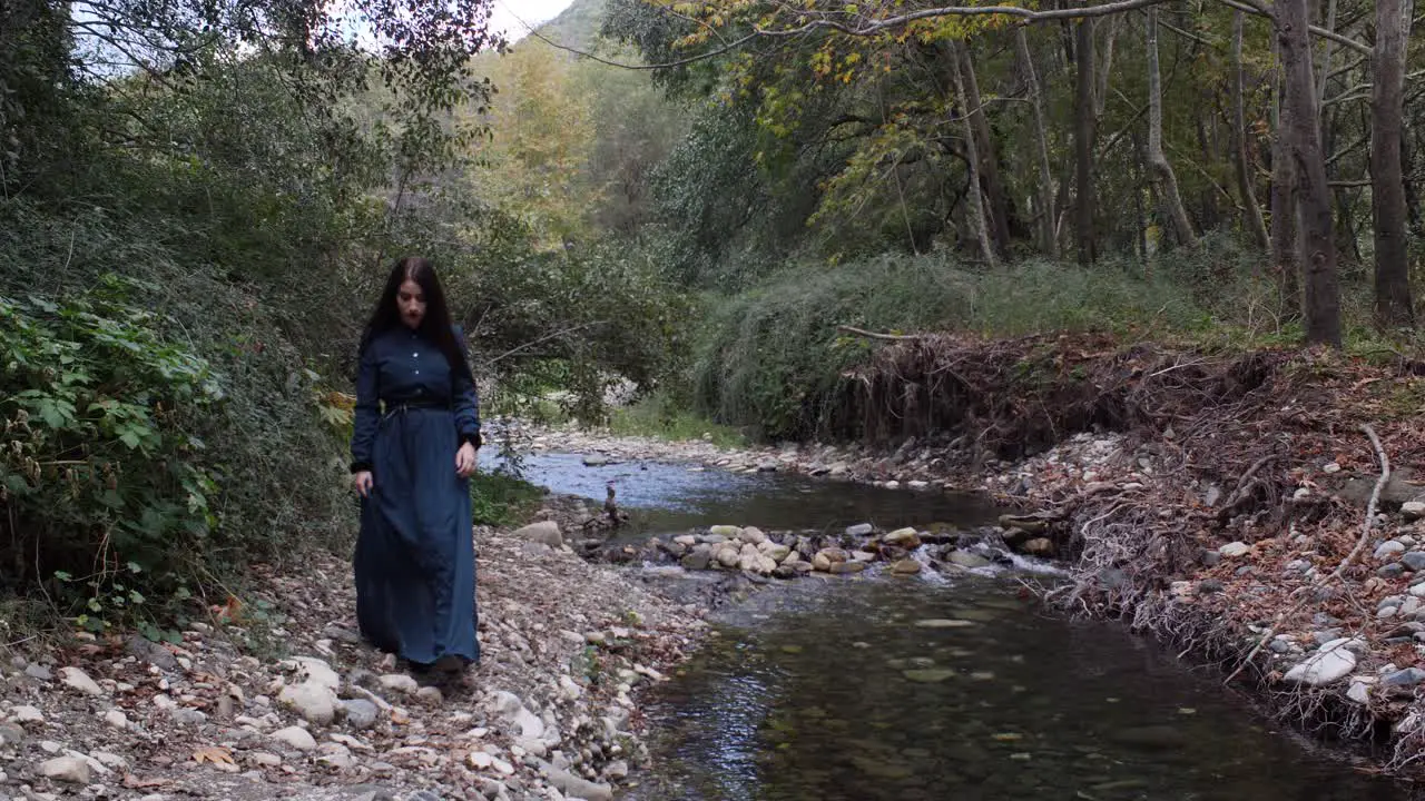 A beautiful woman in a vintage gothic dress walks alone beside a river in the woods