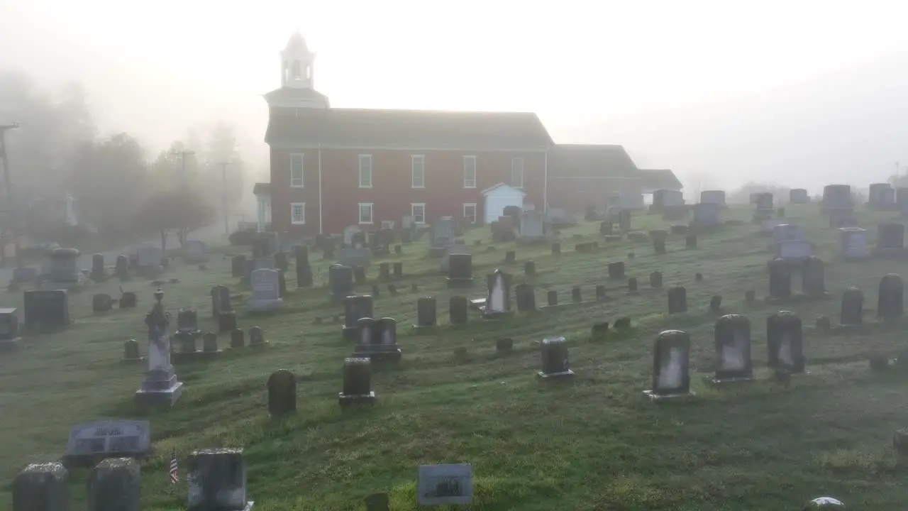 Eerie fog envelops old graveyard in front of red church in establishing shot