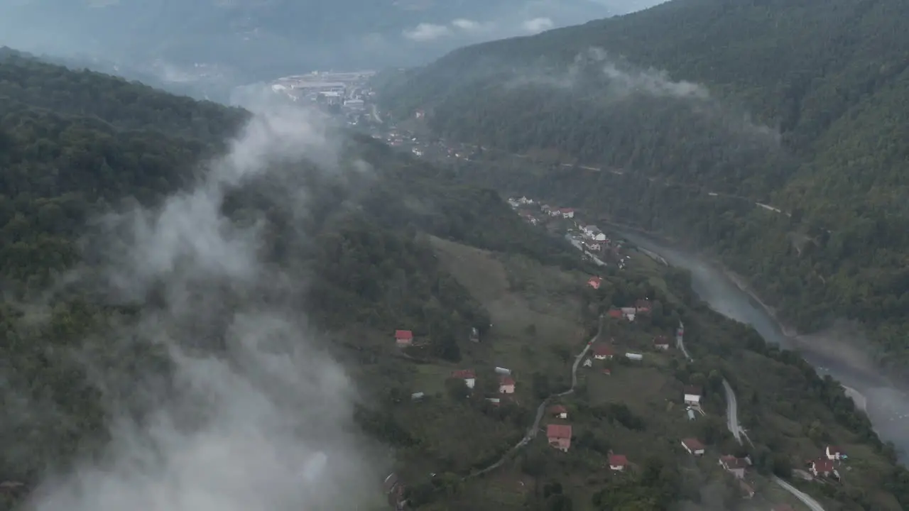 Fly over clouds river stream houses and lush dark forest tree in the balkans