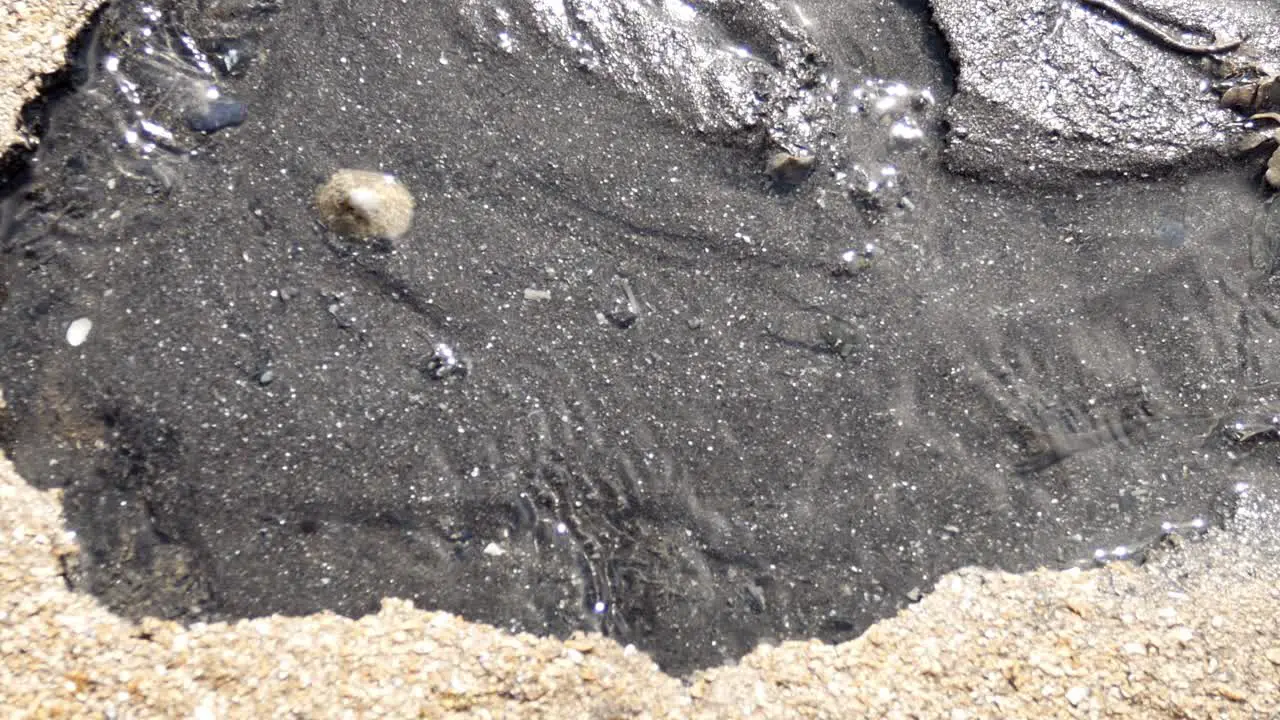 clear flowing water shimmering over black oil polluted textured contrast sand slow dolly right