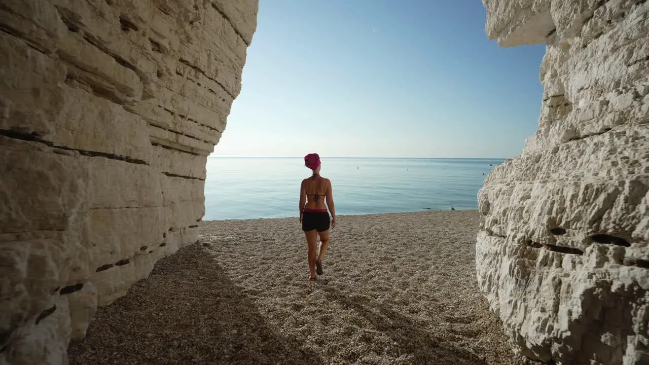 Woman followed from behind walking out of a cave on to the beach towards the blue sea in Italy