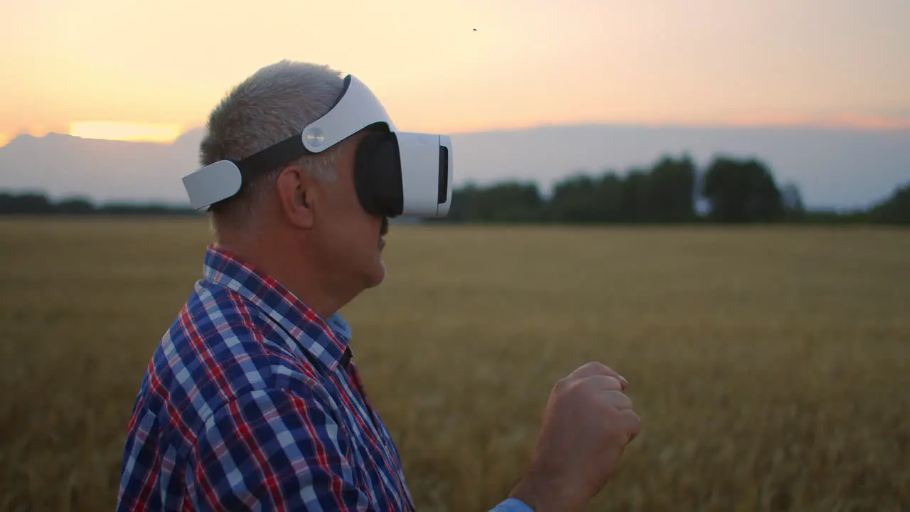 Senior adult farmer in a virtual reality helmet in a field of grain crops In the sunset light an elderly man in a tractor driver uses virtual reality glasses VR technologies and modern agribusiness