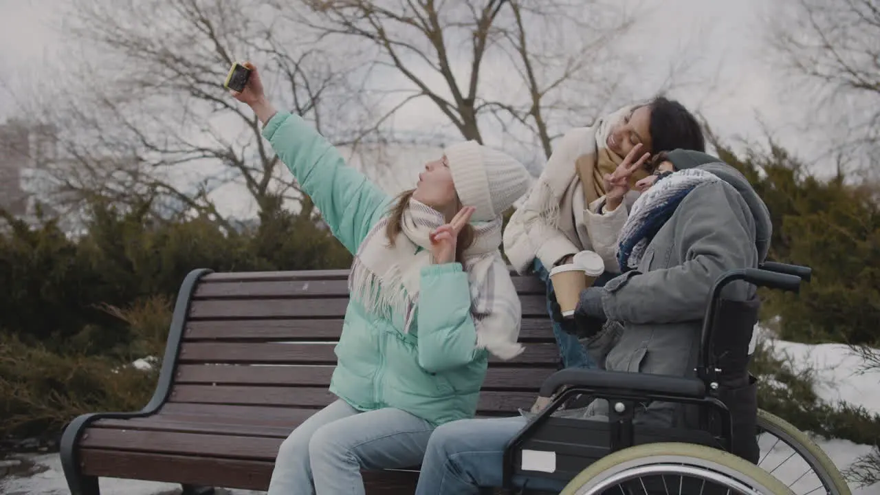 Group Of Friends Two Women And A Disabled Man In Wheelchair Taking A Selfie Video At Urban Park During Winter Season 1