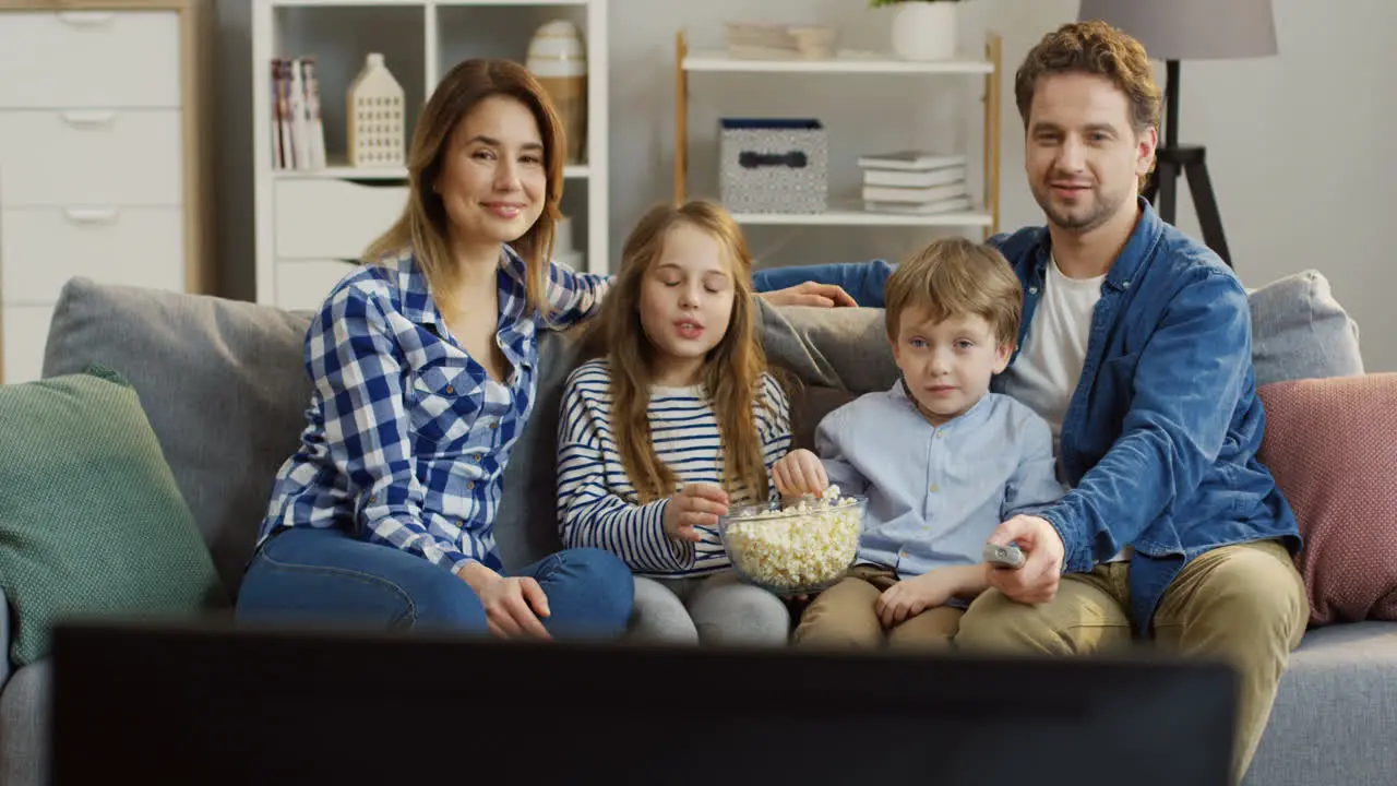 Young Parents With Their Kids Sitting On The Couch In The Living Room Eating Popcorn And Watching Tv