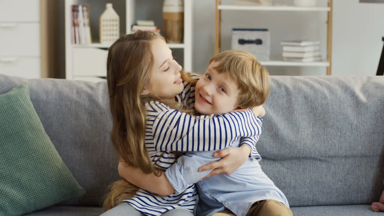 Two Little Cute Children Sister And Brother Sitting On The Sofa With Pillows In The Cozy Room And Hugging Each Other In Front Of The Camera