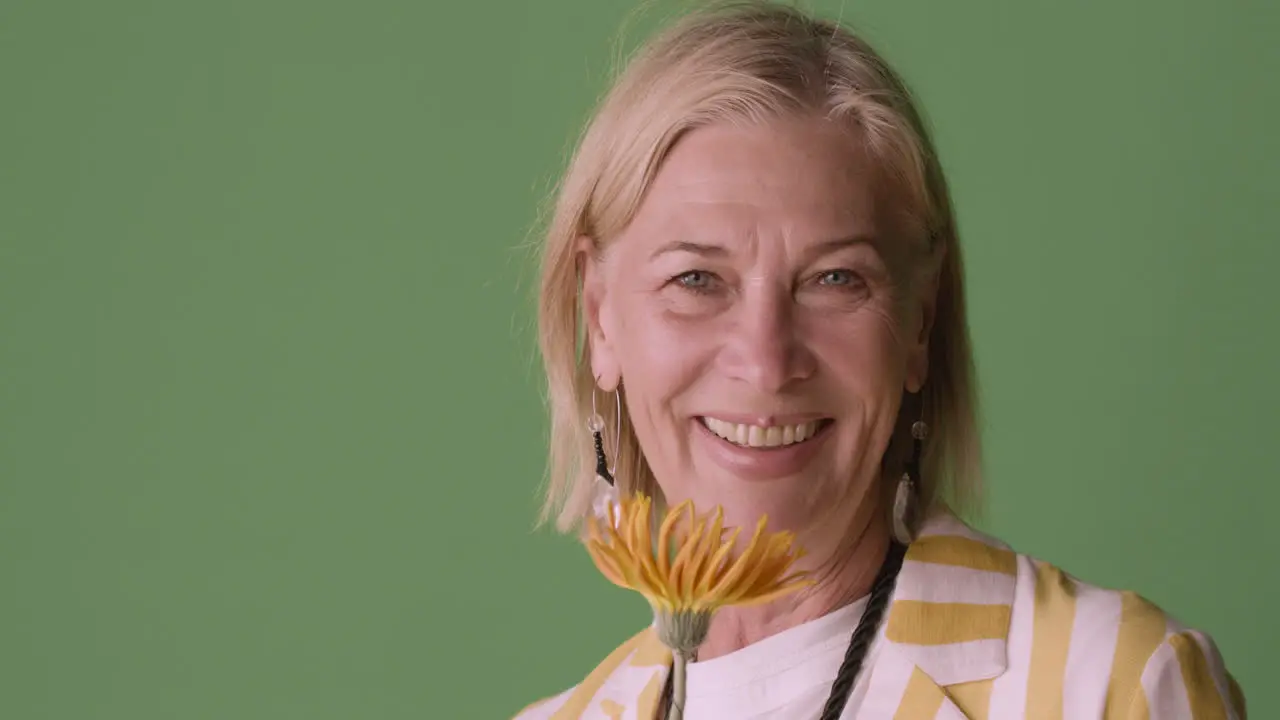 Blonde Mature Woman With Blue Eyes Dressed In Striped Shirt And Accessories Posing Smelling A Sunflower And Smiling At Camera On Green Background