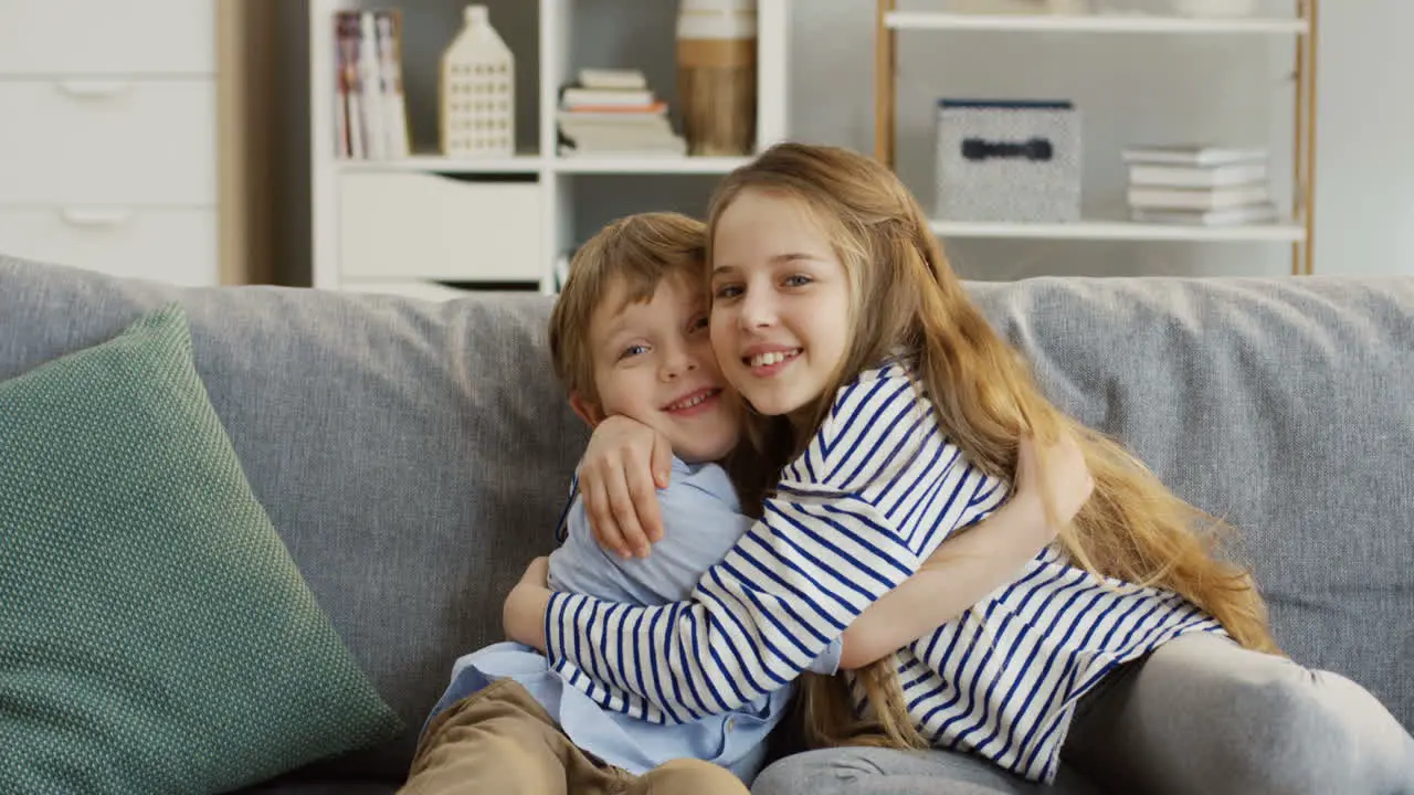 Portrait Shot Of The Cute Small Sister And Brother Sitting On The Couch In The Living Room Hugging Each Other And Posing In Front Of The Camera