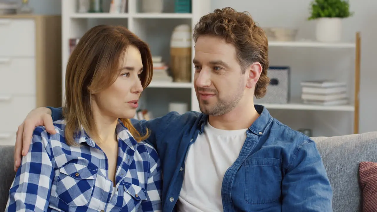 Close Up Of The Young Attractive Man And Woman Sitting Close On The Sofa And Talking Then Their Small Daughter Running To Them And Hugging