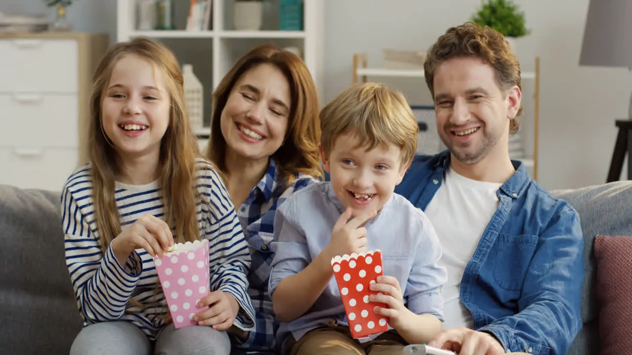 Happy Smiled Family With Popcorn Sitting On The Sofa In The Living Room And Laughing As Watching A Comedy Movie