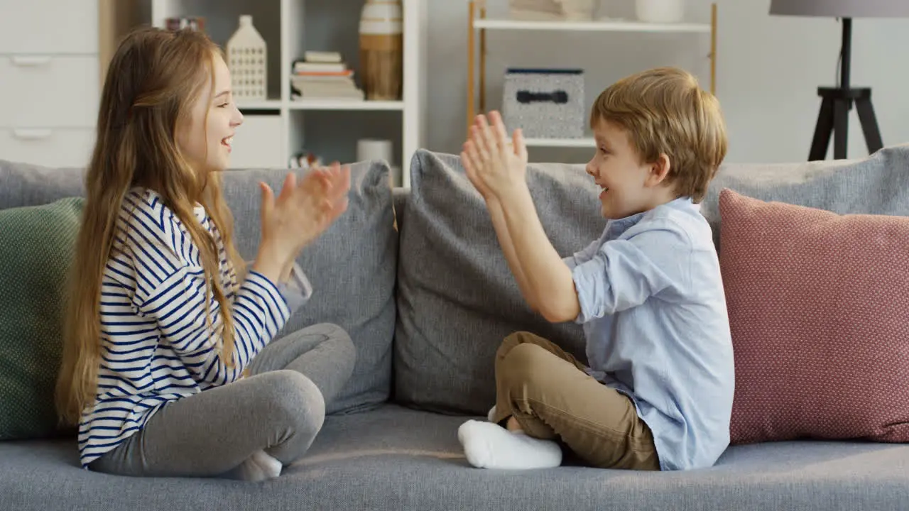 Two Small Cute Kids Sister And Brother Sitting On The Couch With Pillows In The Cozy Room And Playing A Game With Their Hands By Clapping Them