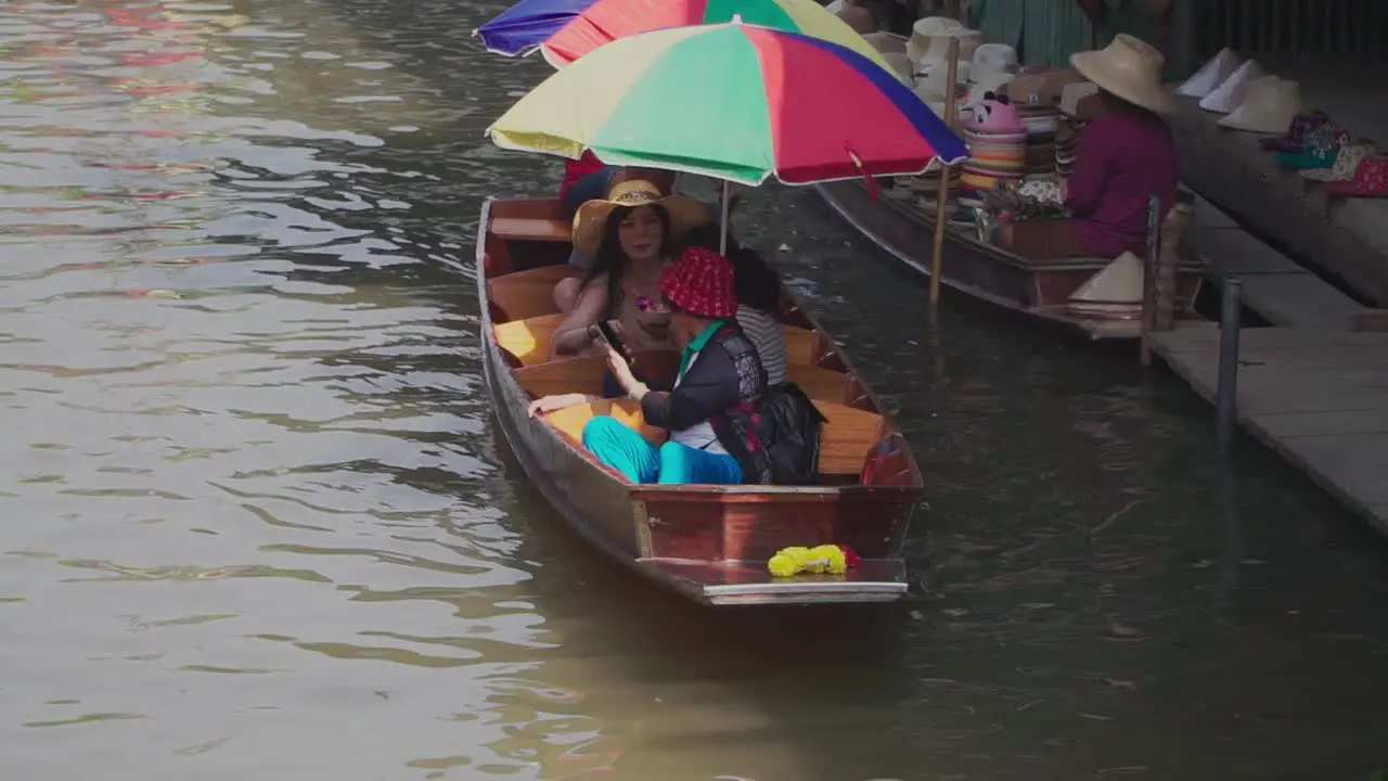 Establishing Shot Man is rowing a boat with tourist in Thailand Floating Market Assorted Hats are displayed on the boat of a vendor in the background