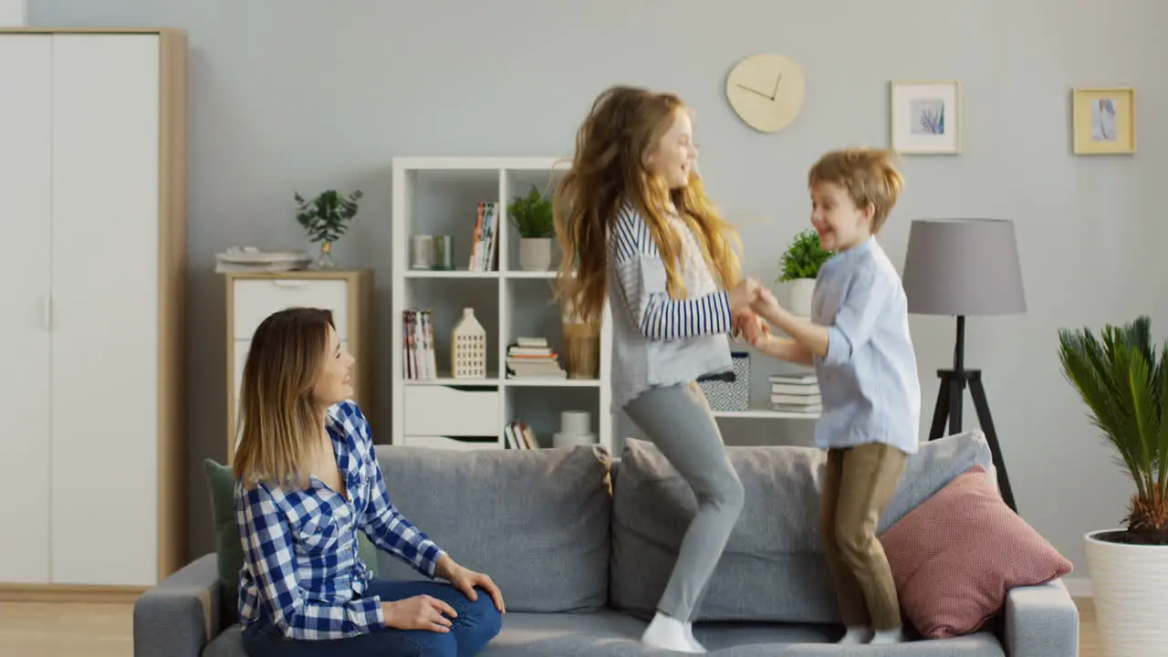 Small Cute Children Brother And Sister Having Fun In The Living Room While Jumping On The Sofa And Their Mother Watching Them