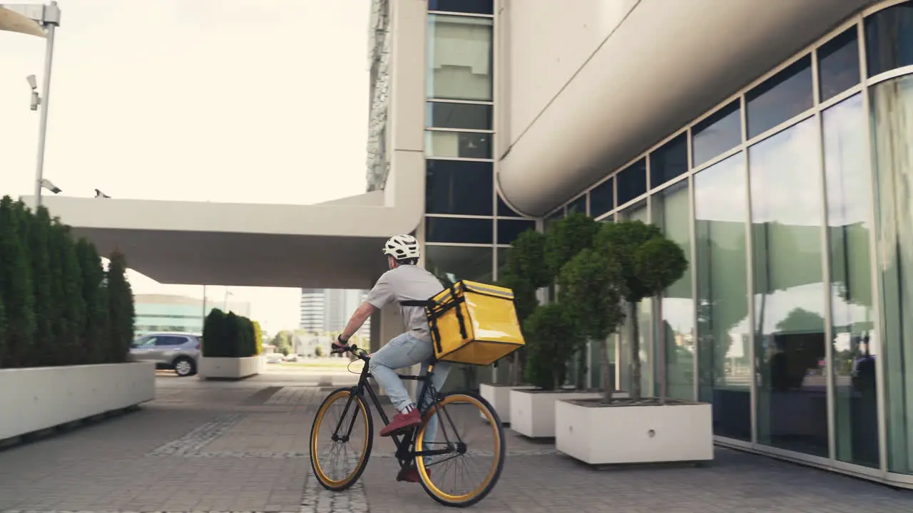 Food Delivery Courier Wearing Thermal Backpack Rides A Bike Next To An Office Building To Deliver Orders For Clients And Customers