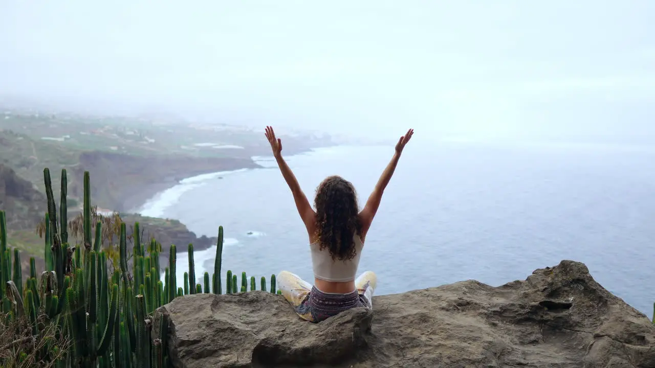 On an island's mountain a young woman practices yoga sitting on a rock meditating in Lotus position overlooking the ocean