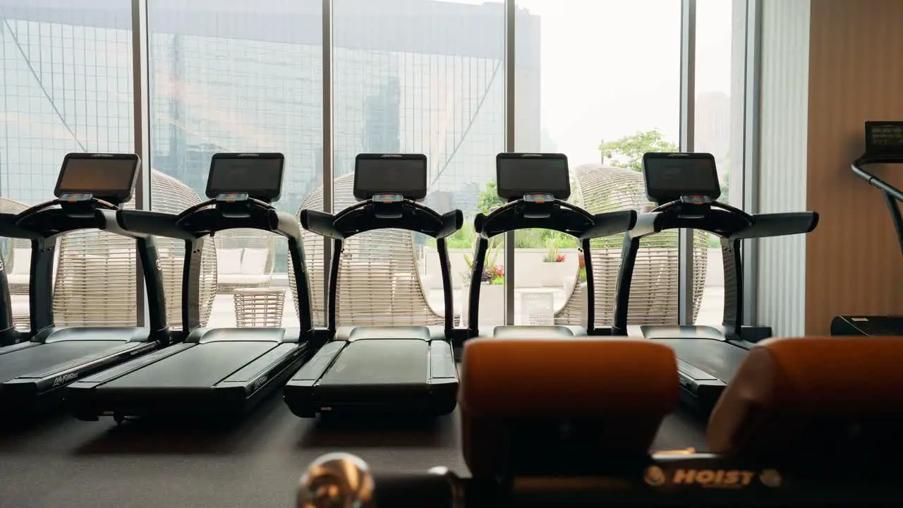 Panning across multiple treadmills in a fitness center situated in front of expansive windows that provide a view to the outdoors