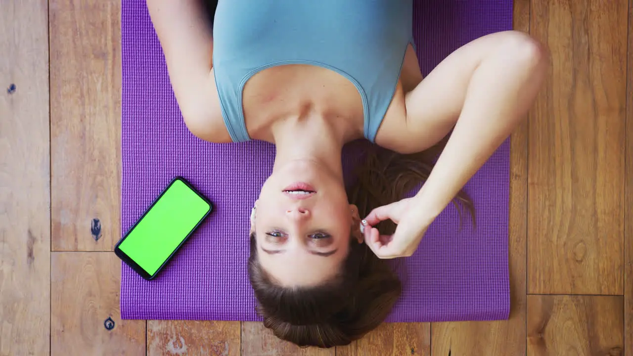 Overhead View Of Woman Lying On Exercise Mat At Home Taking Out Wireless Earphones After Workout