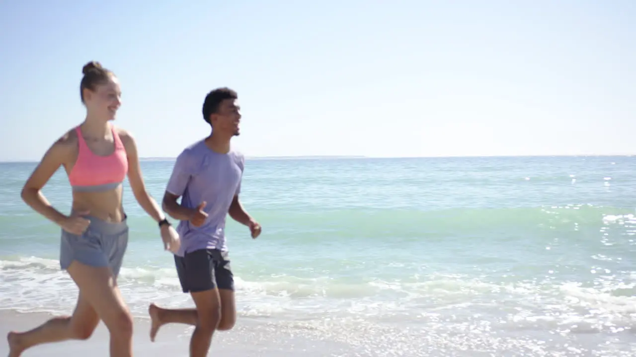 A young Caucasian woman and a biracial man dressed in athletic wear are jogging along the beach