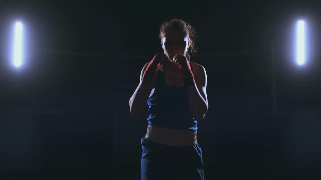 A beautiful female boxer strikes directly into the camera looking into the camera and moving forward on a dark background with a backlight Steadicam shot