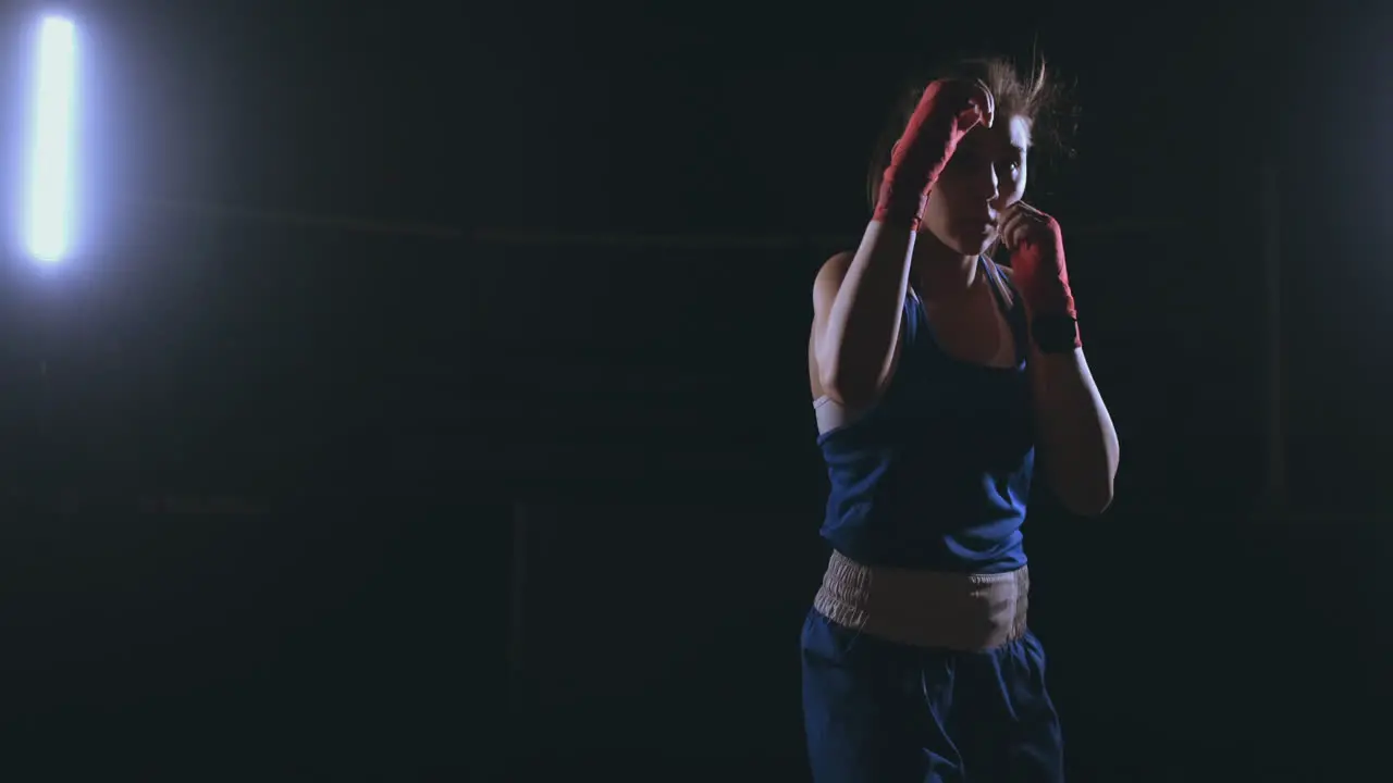 Looking into the camera a beautiful female boxer strikes against a dark background with a backlit light Steadicam shot