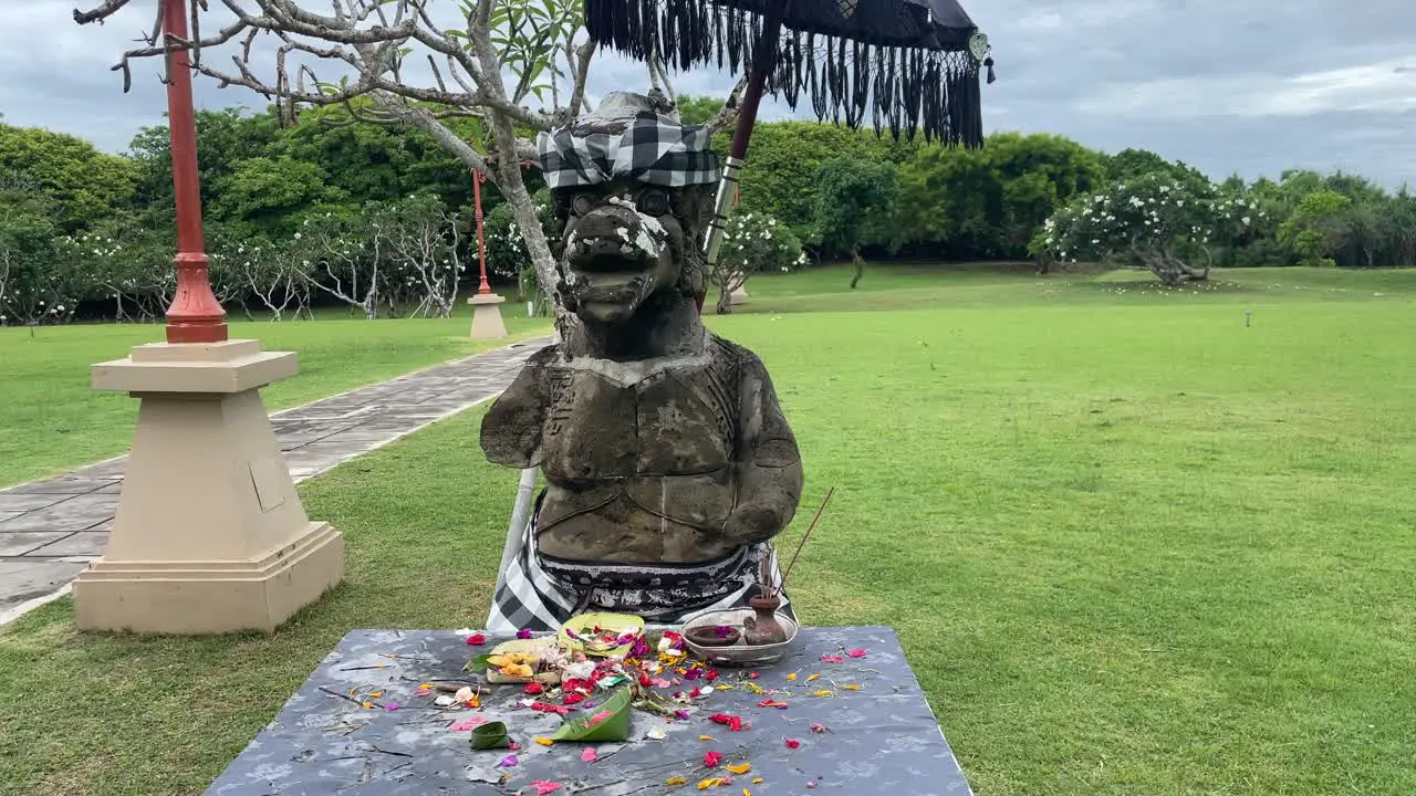 Tilt up to a stone God statue flowers and offerings in front of the figure in a green lush park