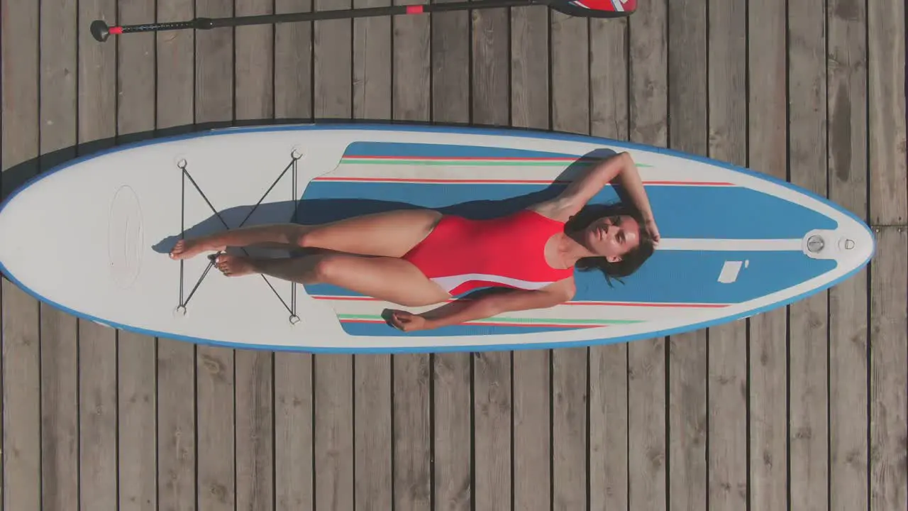 Aerial View Of A Girl In Red Swimsuit Lying On The Surfboard With A Paddle Next To Her On The Wooden Harbor 1