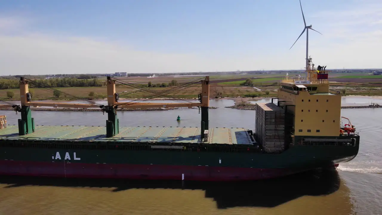 Aerial Tracking Shot Of Port Side View Of Aal Paris Cargo Ship Along Oude Maas With Still Wind Turbine In Background