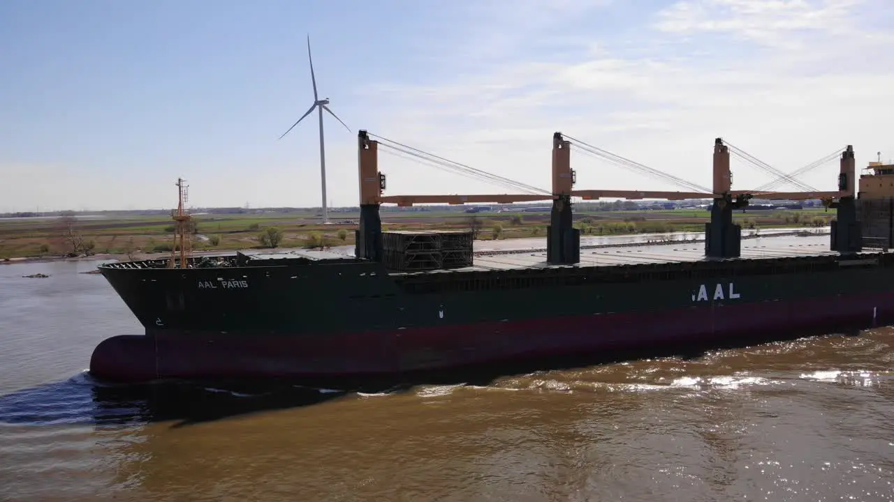 Aerial Port Side View Of Aal Paris Cargo Ship Travelling Along Oude Maas With Still Wind Turbine In Background