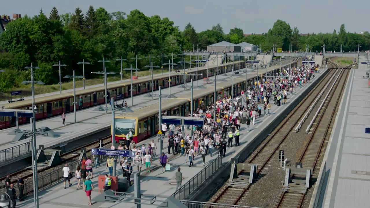 An outdoor scene depicting a bustling train station with multiple tracks and platforms featuring a crowd of people trains and encircled by trees and buildings